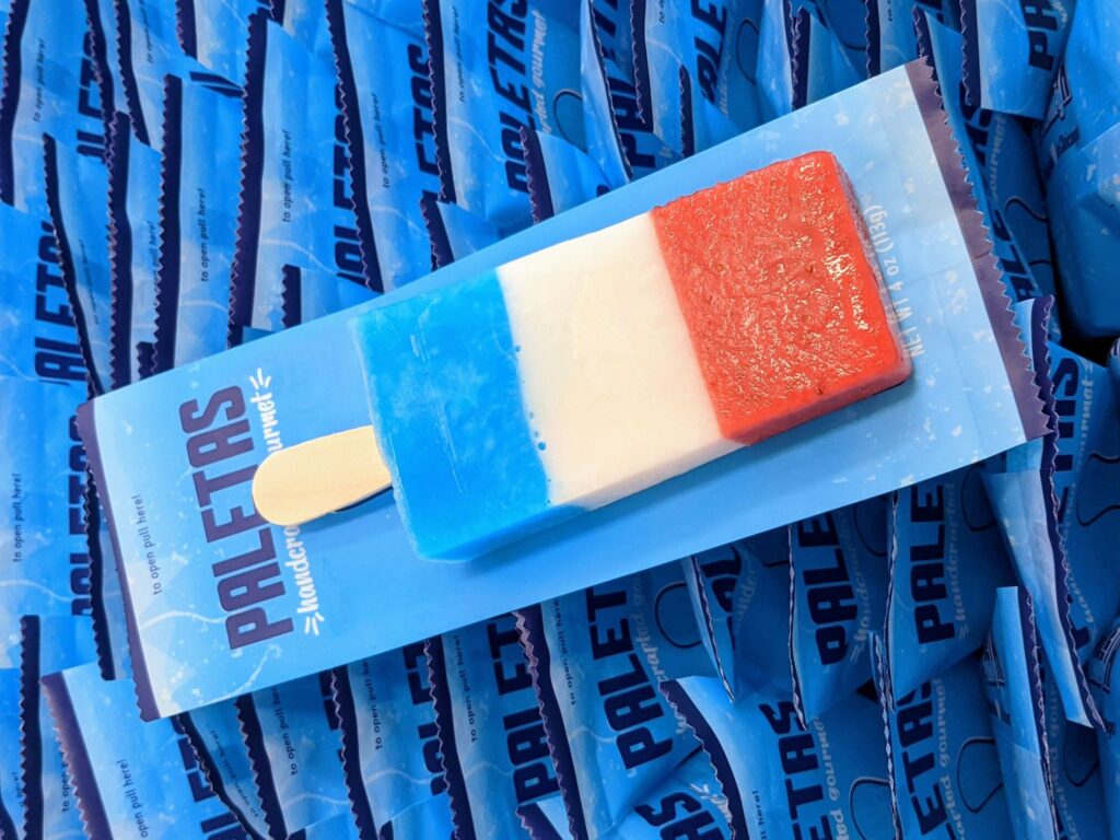 Close-up of Paleta Factory's red, white, and blue popsicle, representing strawberry, lime, and blue raspberry flavors, packaged in a blue wrapper.