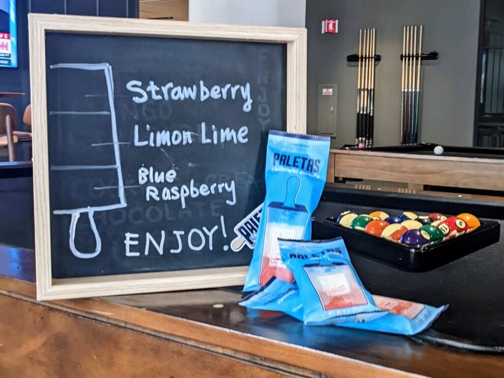 Chalkboard sign displaying Paleta Factory's custom flavors: strawberry, lime, and blue raspberry, with wrapped popsicles placed on the table.